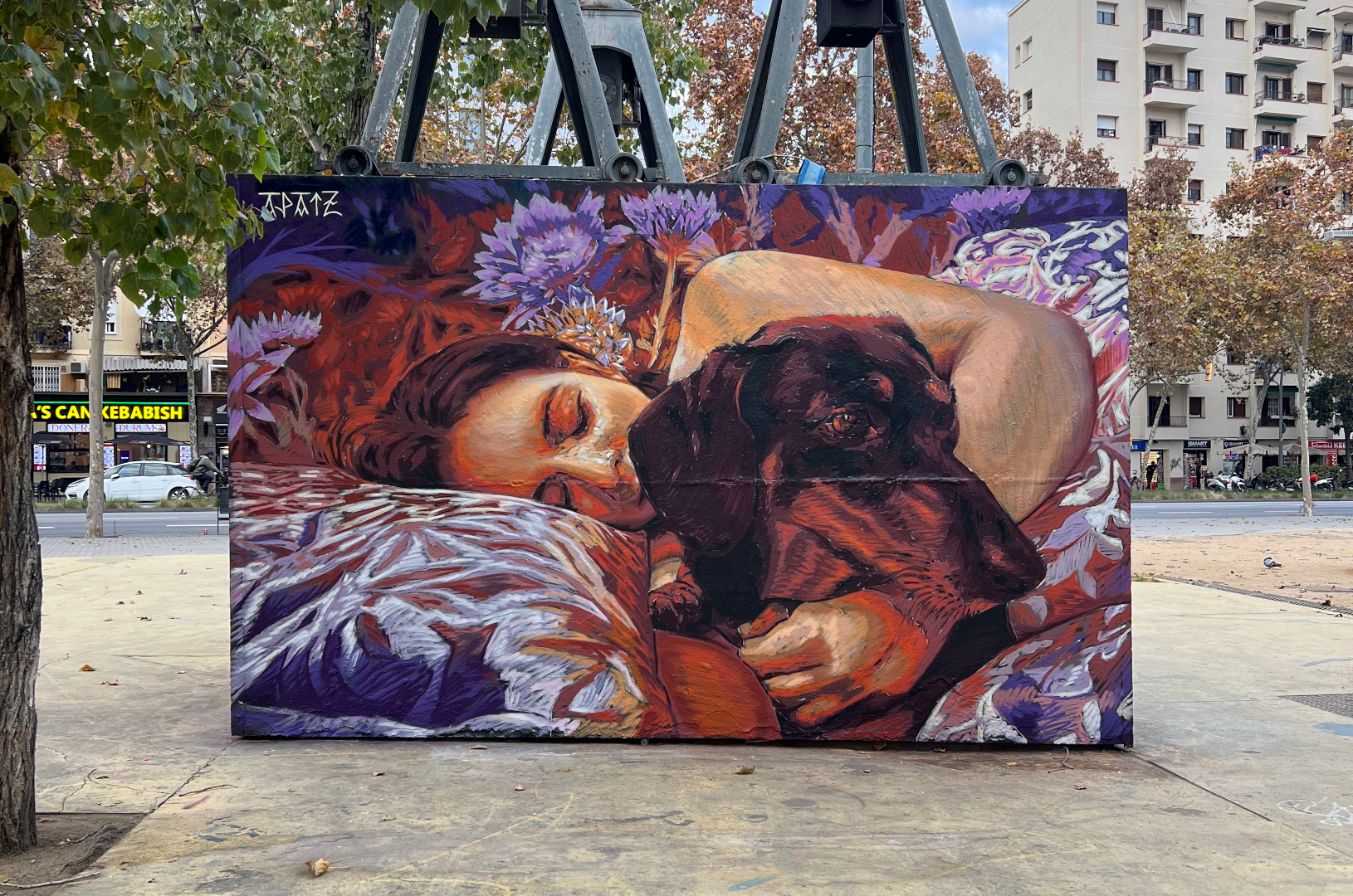 Fresque murale représentant une personne allongée, les yeux fermés, enlaçant un chien noir endormi. La scène, peinte dans des tons chauds de rouge, violet et orange, évoque la tendresse et l’intimité. L’œuvre est installée sur la place des Trois Cheminées (Tres Xemeneies) à Barcelone, dans un environnement urbain marqué par des structures industrielles et des arbres en arrière-plan.