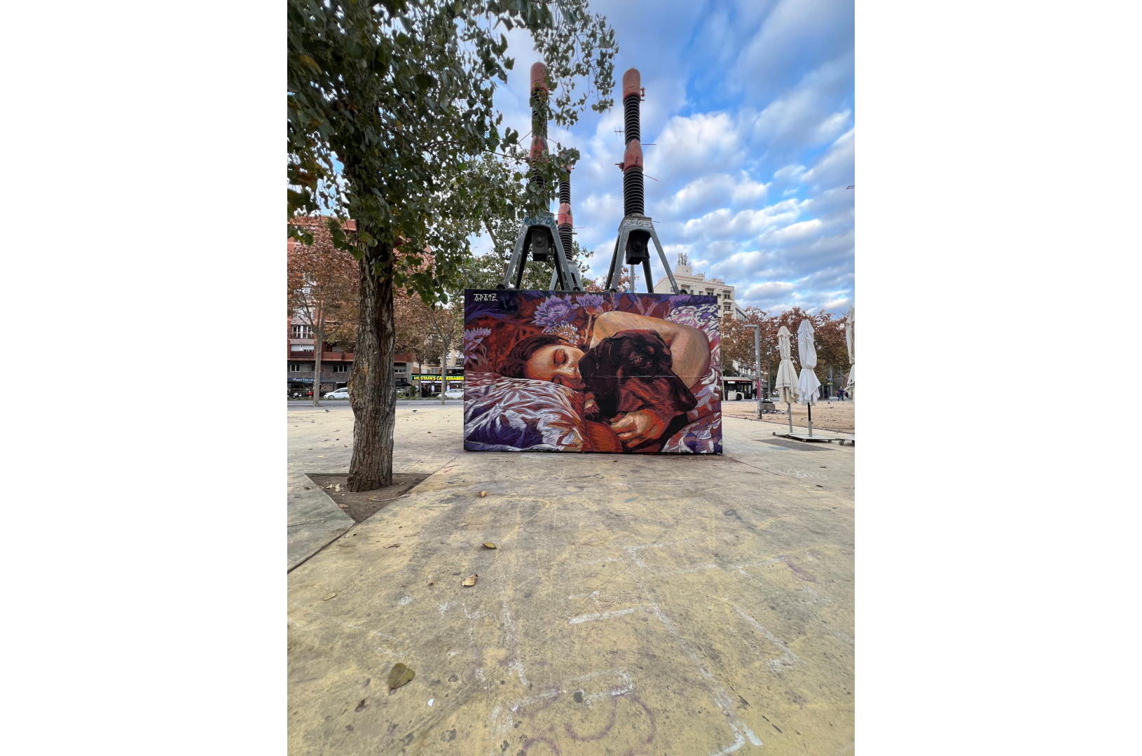 Fresque murale représentant une personne allongée, les yeux fermés, enlaçant un chien noir endormi. La scène, peinte dans des tons chauds de rouge, violet et orange, évoque la tendresse et l’intimité. L’œuvre est installée sur la place des Trois Cheminées (Tres Xemeneies) à Barcelone, dans un environnement urbain marqué par des structures industrielles et des arbres en arrière-plan.