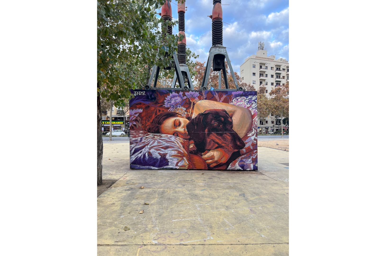 Fresque murale représentant une personne allongée, les yeux fermés, enlaçant un chien noir endormi. La scène, peinte dans des tons chauds de rouge, violet et orange, évoque la tendresse et l’intimité. L’œuvre est installée sur la place des Trois Cheminées (Tres Xemeneies) à Barcelone, dans un environnement urbain marqué par des structures industrielles et des arbres en arrière-plan.