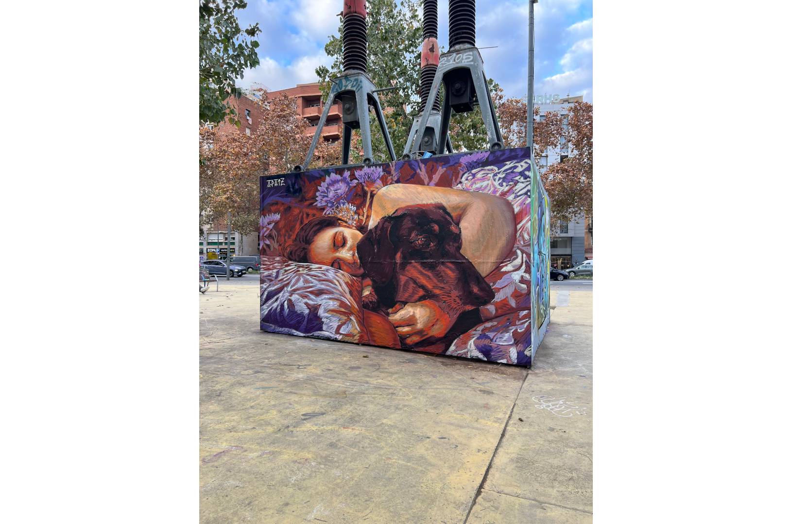 Fresque murale représentant une personne allongée, les yeux fermés, enlaçant un chien noir endormi. La scène, peinte dans des tons chauds de rouge, violet et orange, évoque la tendresse et l’intimité. L’œuvre est installée sur la place des Trois Cheminées (Tres Xemeneies) à Barcelone, dans un environnement urbain marqué par des structures industrielles et des arbres en arrière-plan.