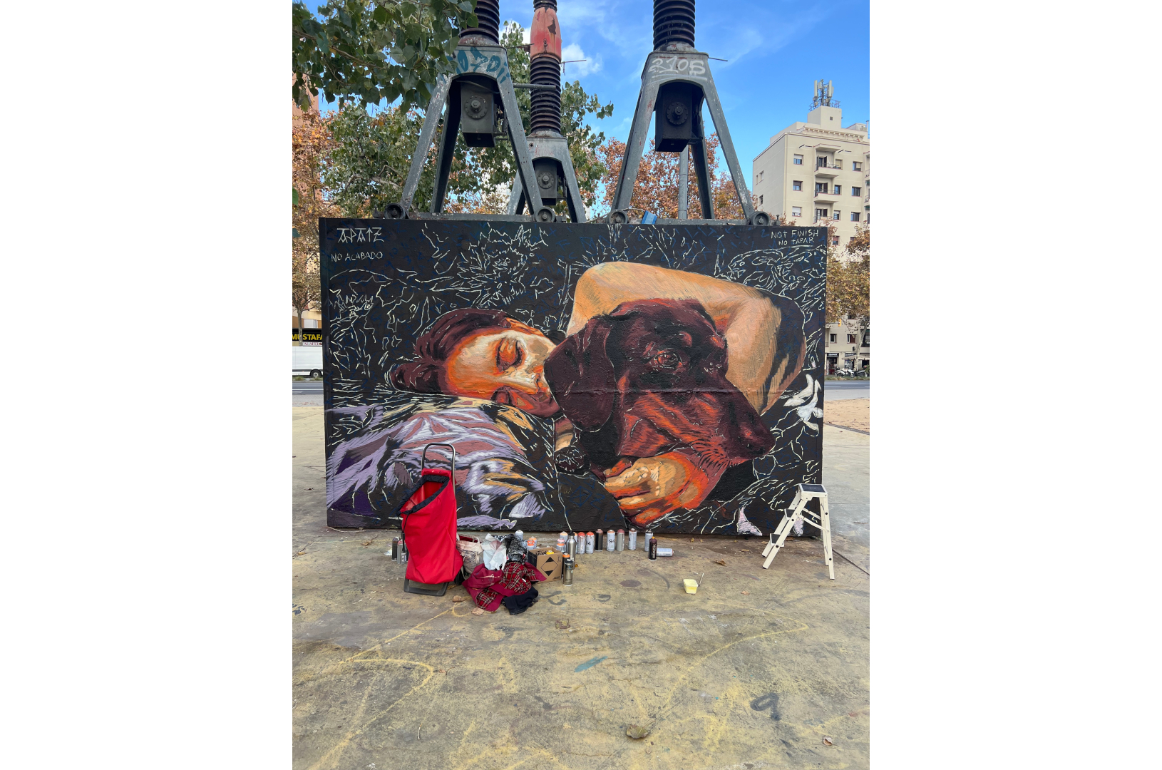 Fresque murale représentant une personne allongée, les yeux fermés, enlaçant un chien noir endormi. La scène, peinte dans des tons chauds de rouge, violet et orange, évoque la tendresse et l’intimité. L’œuvre est installée sur la place des Trois Cheminées (Tres Xemeneies) à Barcelone, dans un environnement urbain marqué par des structures industrielles et des arbres en arrière-plan.